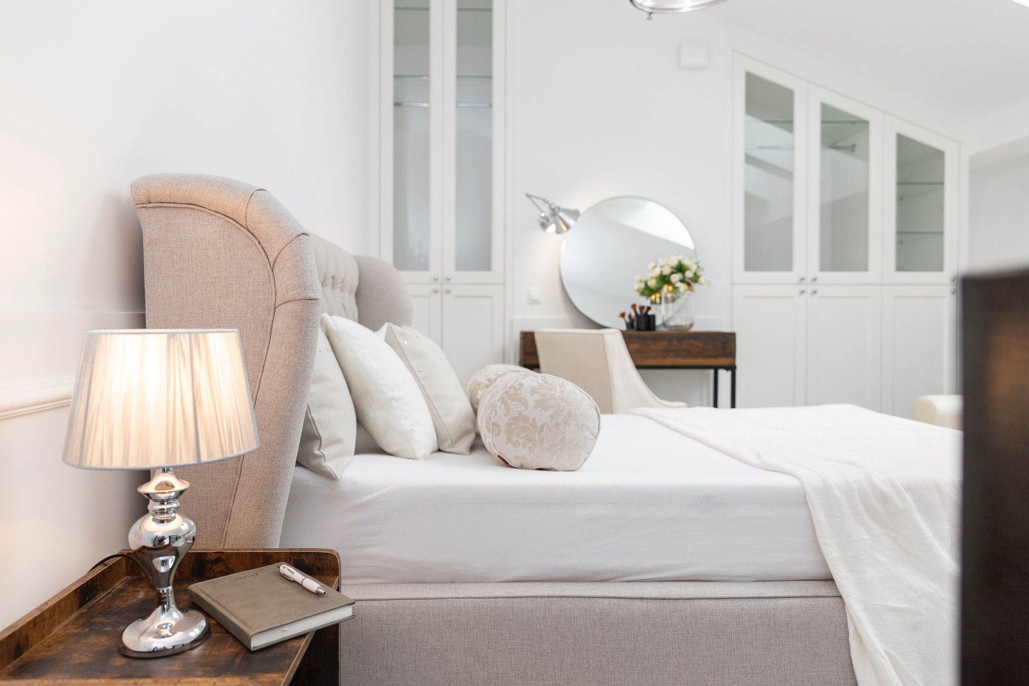 Jasna sypialnia z beżowym łóżkiem, białą pościelą, okrągłym lustrem i drewnianą szafką nocną z lampką. W tle biała zabudowa szaf z przeszklonymi frontami.