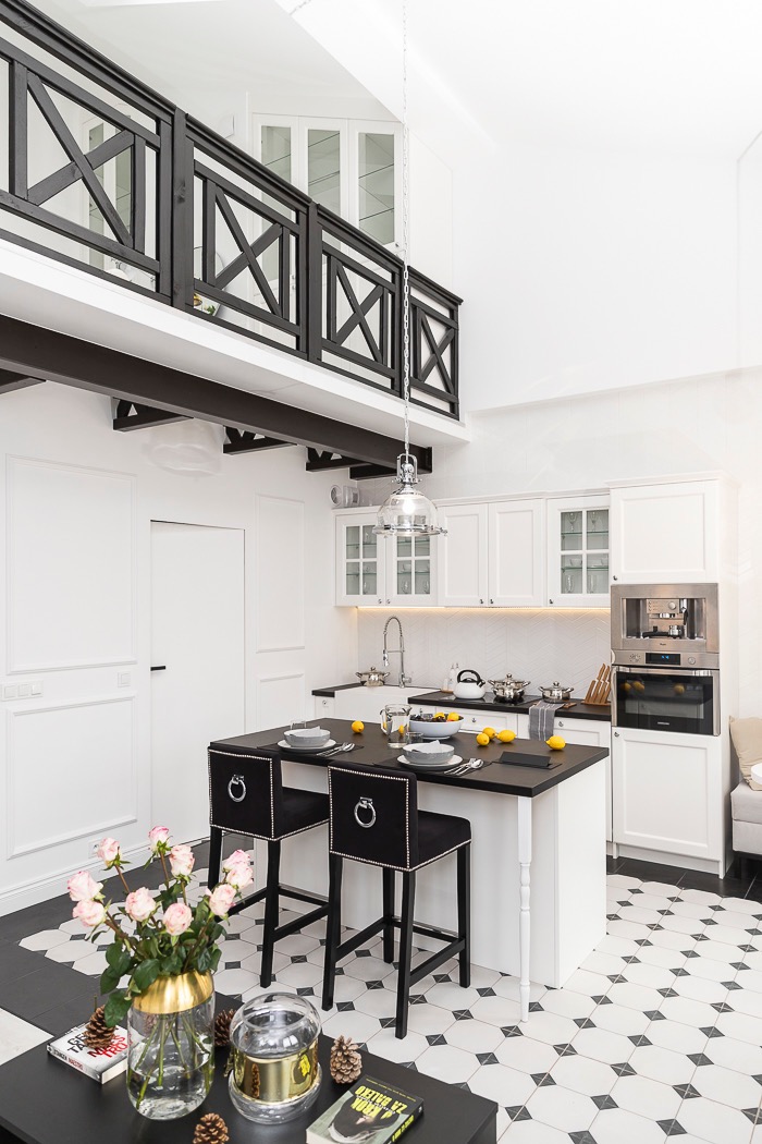 Wysoka, jasna kuchnia z antresolą. Białe szafki, czarny blat, wysokie krzesła barowe z ozdobnymi pierścieniami. Podłoga w czarno-białą szachownicę.