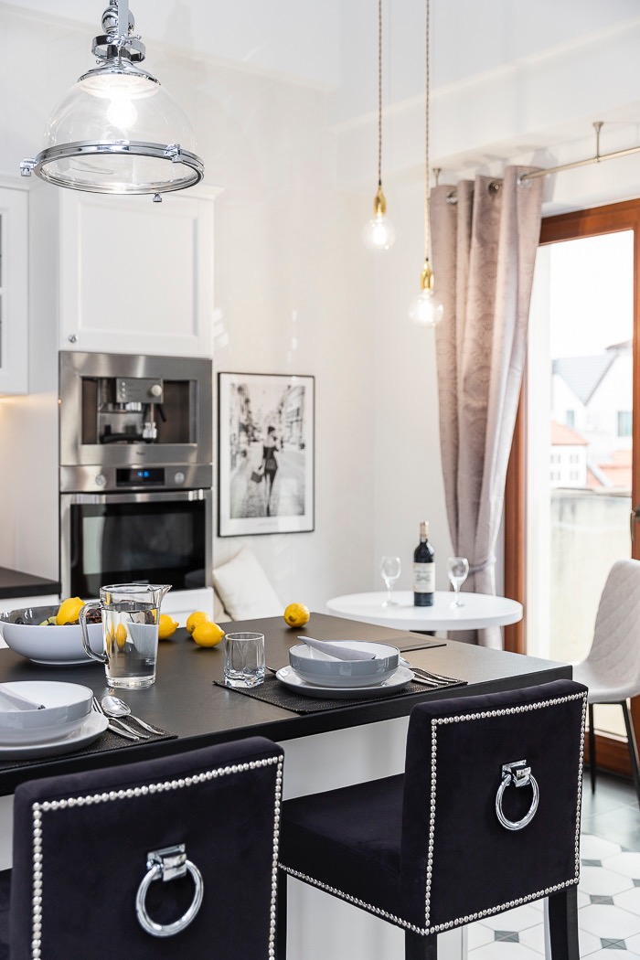 Elegancka kuchnia z czarnym blatem, owocami i stylowymi krzesłami. W tle zabudowa kuchenna z piekarnikiem i ekspresem do kawy. Całość dopełniają lampy wiszące.