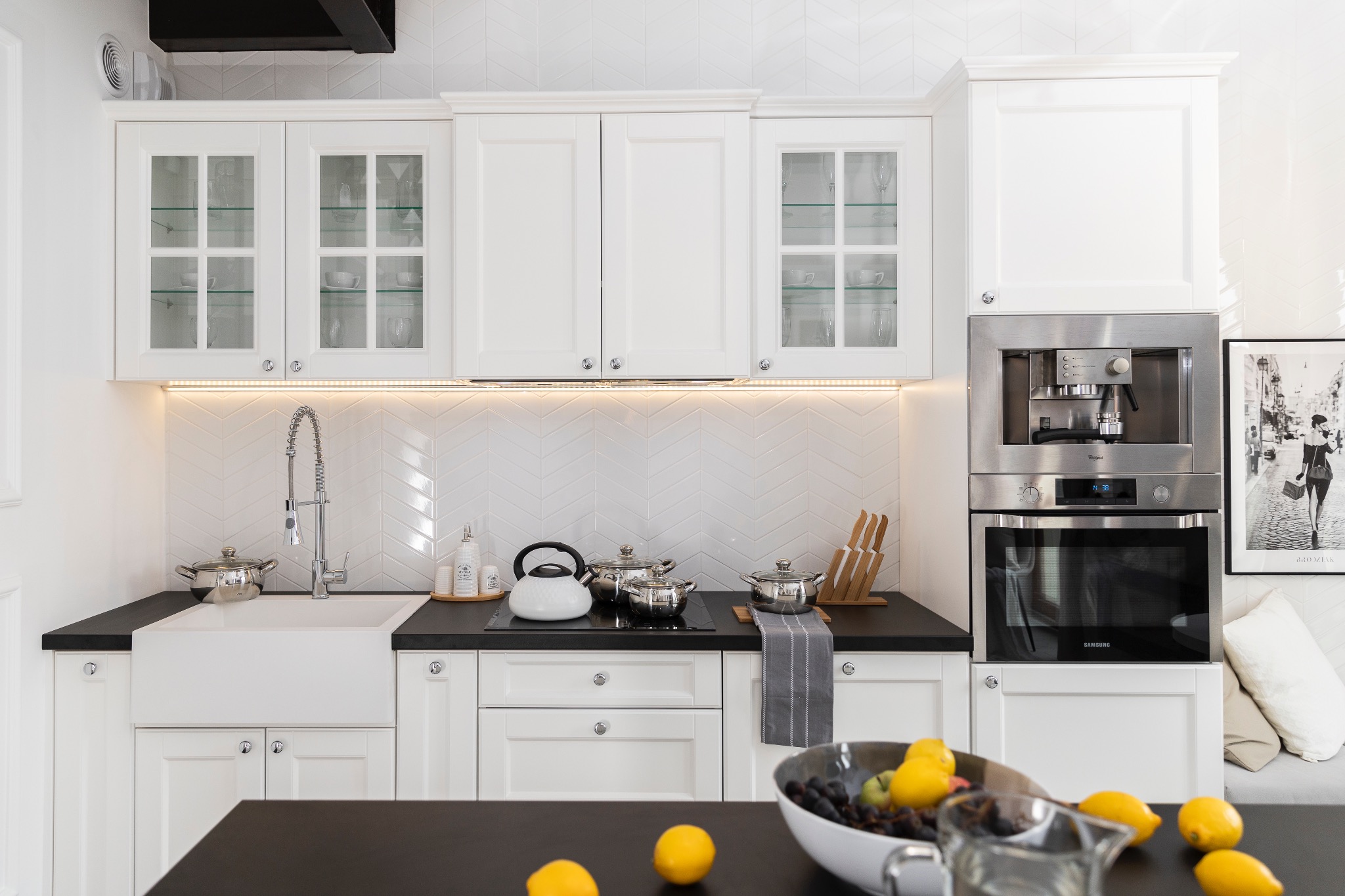 Jasna kuchnia w stylu klasycznym z białymi szafkami, czarnym blatem i sprzętem AGD w zabudowie. Uwagę przykuwają płytki ułożone w jodełkę na ścianie.