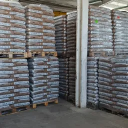 Składowane palety z workami pelletu marki Igła 15kg, owinięte folią stretch, w magazynie. Widoczne drewniane elementy konstrukcyjne budynku.