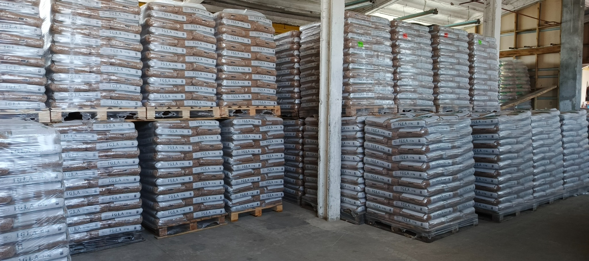 Składowane palety z workami pelletu marki Igła 15kg, owinięte folią stretch, w magazynie. Widoczne drewniane elementy konstrukcyjne budynku.