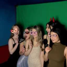 Grupa pięciu uśmiechniętych, młodych kobiet pozuje z rekwizytami (maski, okulary) na tle zielonego ekranu w fotobudce. Jedna z nich ma rogi diabła.