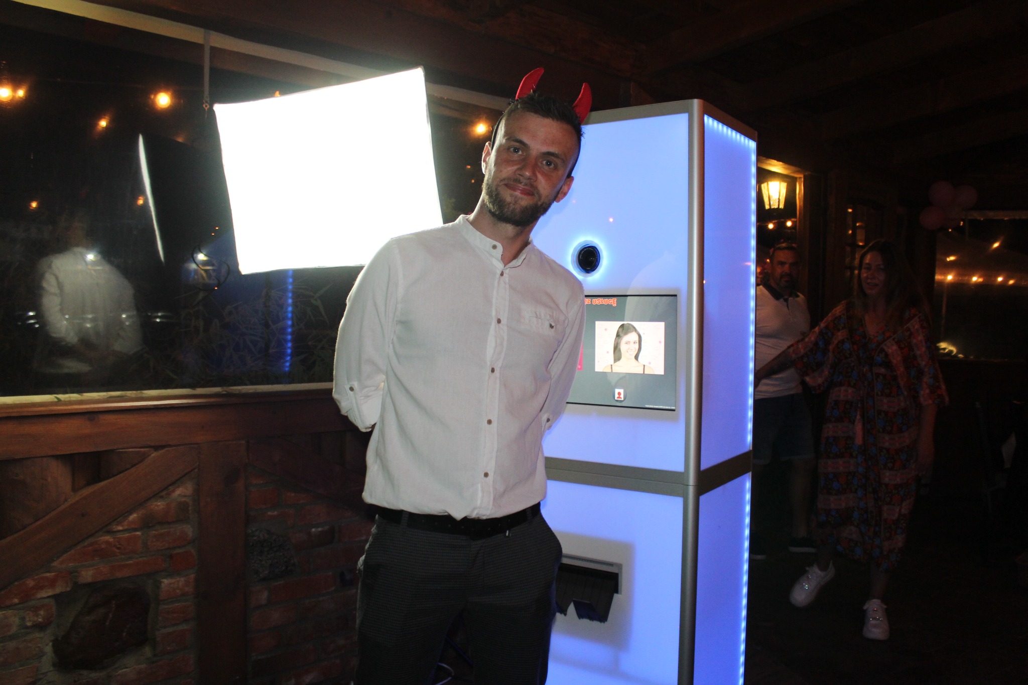 Mężczyzna z rogami diabła pozuje przy fotobudce z niebieskim podświetleniem LED. Na ekranie widoczna twarz kobiety. W tle para i dekoracje.