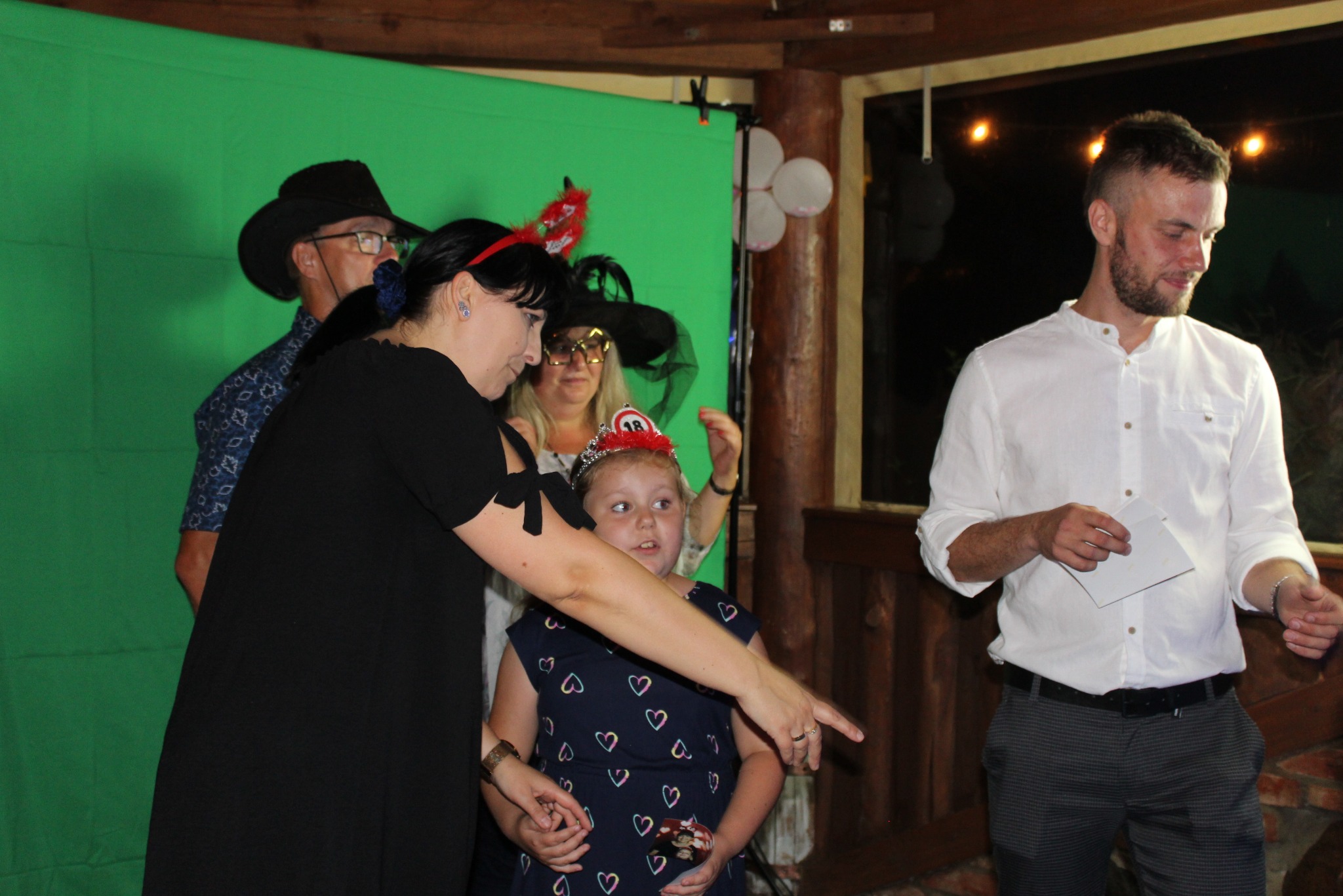 Uczestnicy imprezy pozują na tle zielonego ekranu, dekoracje balonowe w tle. Kobieta w czarnej sukience wskazuje palcem na ekran. Mężczyzna w białej koszuli trzyma kartkę.