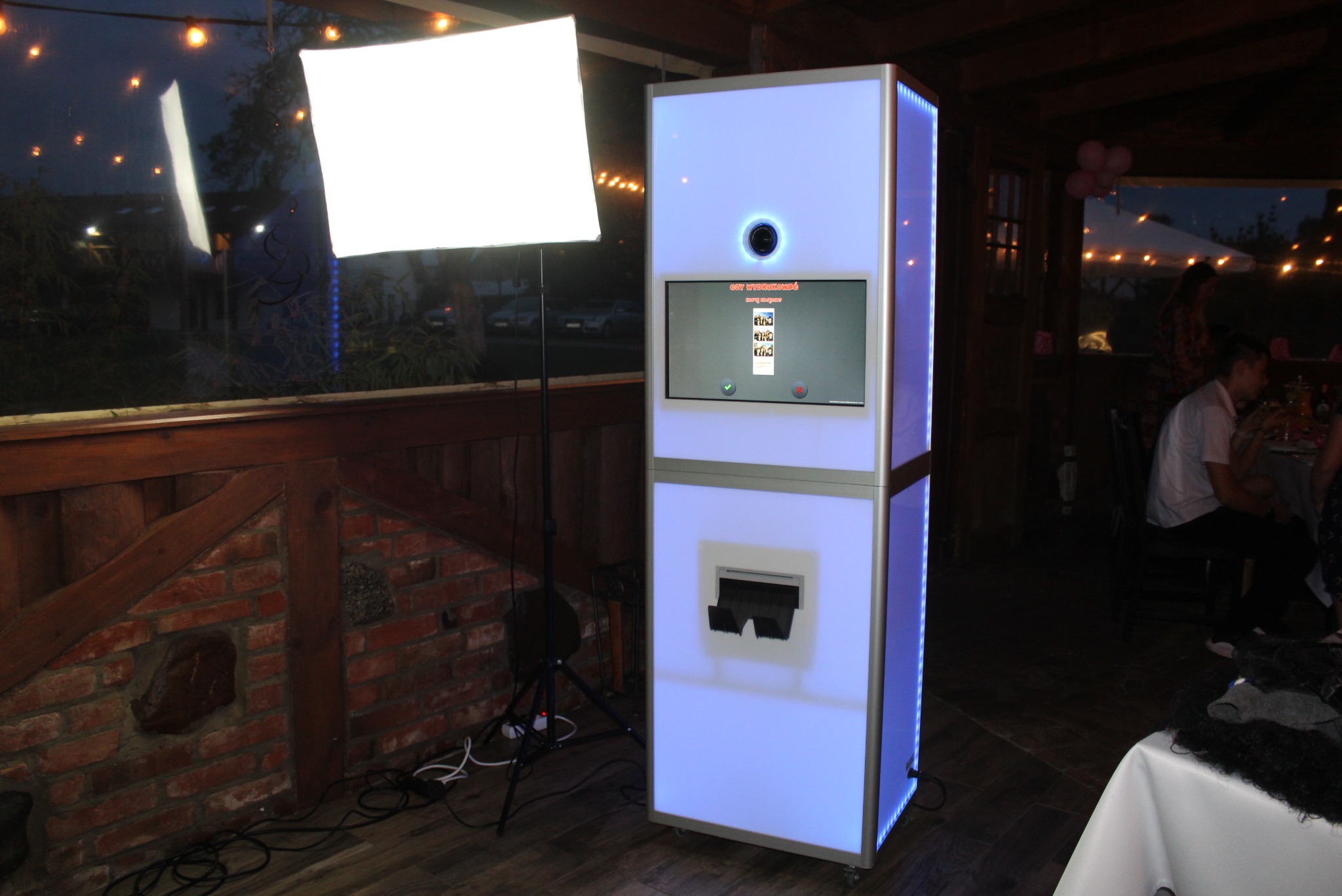 Podświetlana fotobudka z ekranem dotykowym na przyjęciu, widoczne osoby przy stole w tle, oświetlenie dekoracyjne i ceglana ściana w rustykalnym stylu.