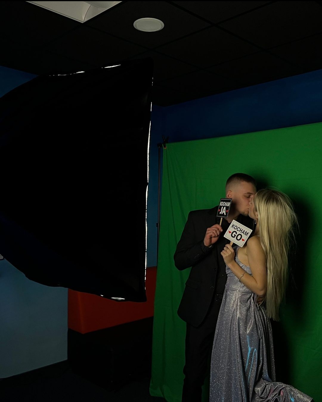 Para całuje się w fotobudce na tle zielonego ekranu, trzymając rekwizyty z napisem 'Kocham Ja' i 'Kocham Go', oświetlenie studyjne po lewej.