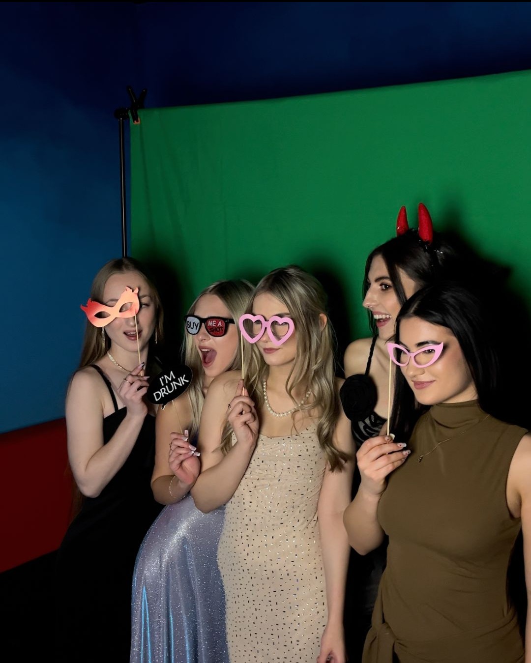 Grupa pięciu uśmiechniętych, młodych kobiet pozuje z rekwizytami (maski, okulary) na tle zielonego ekranu w fotobudce. Jedna z nich ma rogi diabła.