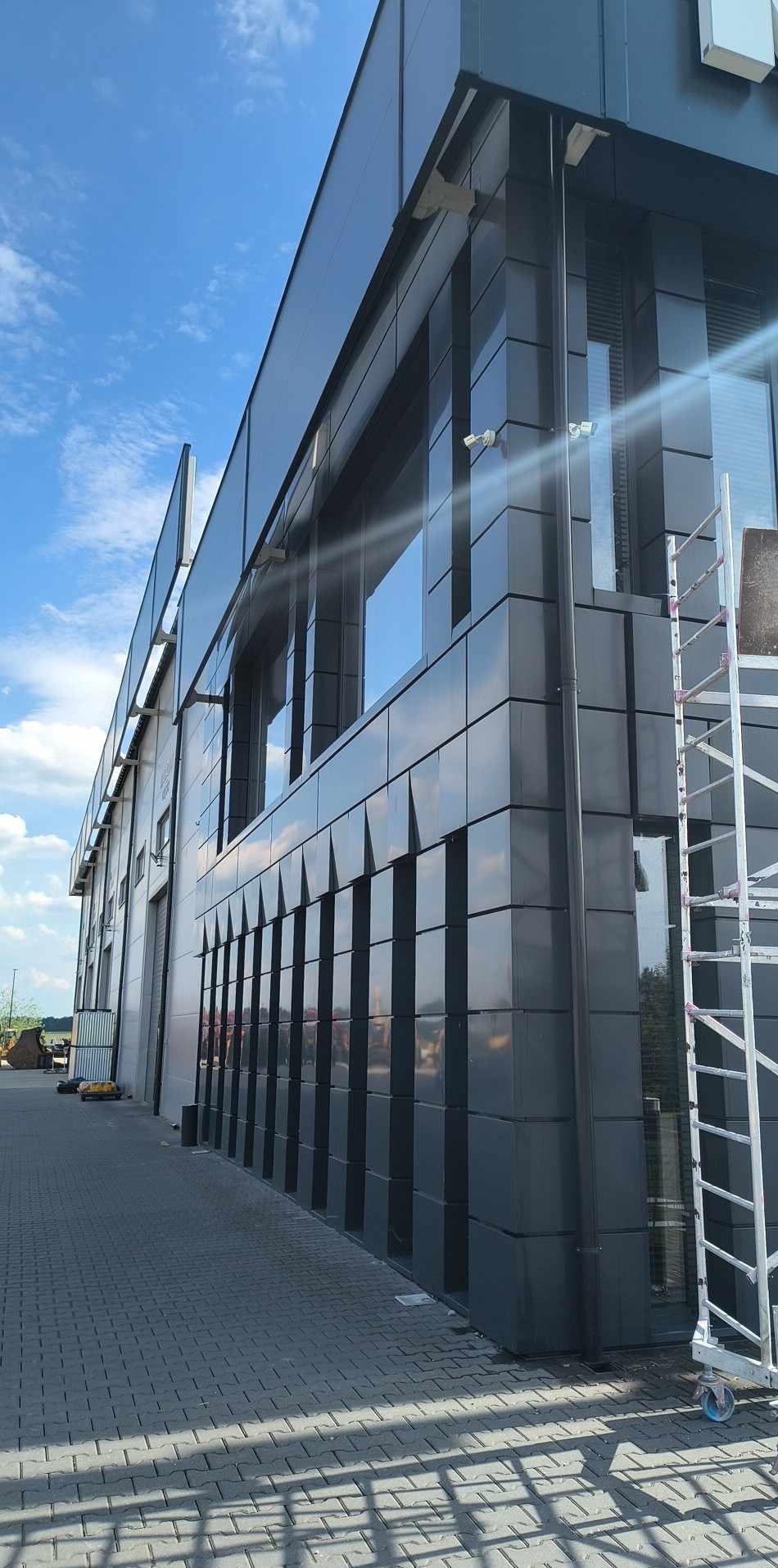 Nowoczesny budynek biurowy z ciemną elewacją i dużymi oknami, częściowo widoczna drabina. Kostka brukowa przed budynkiem, błękitne niebo w tle.