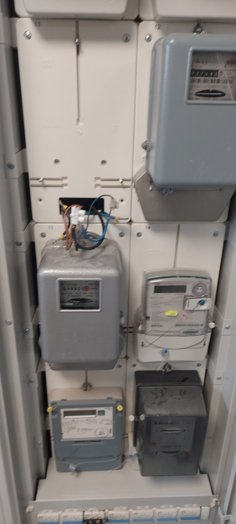 Szafa elektryczna z zamontowanymi różnymi licznikami energii elektrycznej, widoczne przewody i bezpieczniki.