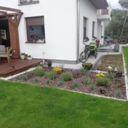 Estetyczny ogród przydomowy z drewnianym tarasem, rabatami kwiatowymi wykończonymi obrzeżem i równo przystrzyżonym trawnikiem.