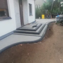 Ułożona kostka brukowa przed domem z trzema schodami, obrzeża w kolorze grafitowym. Widoczne elementy domu i fragment terenu.