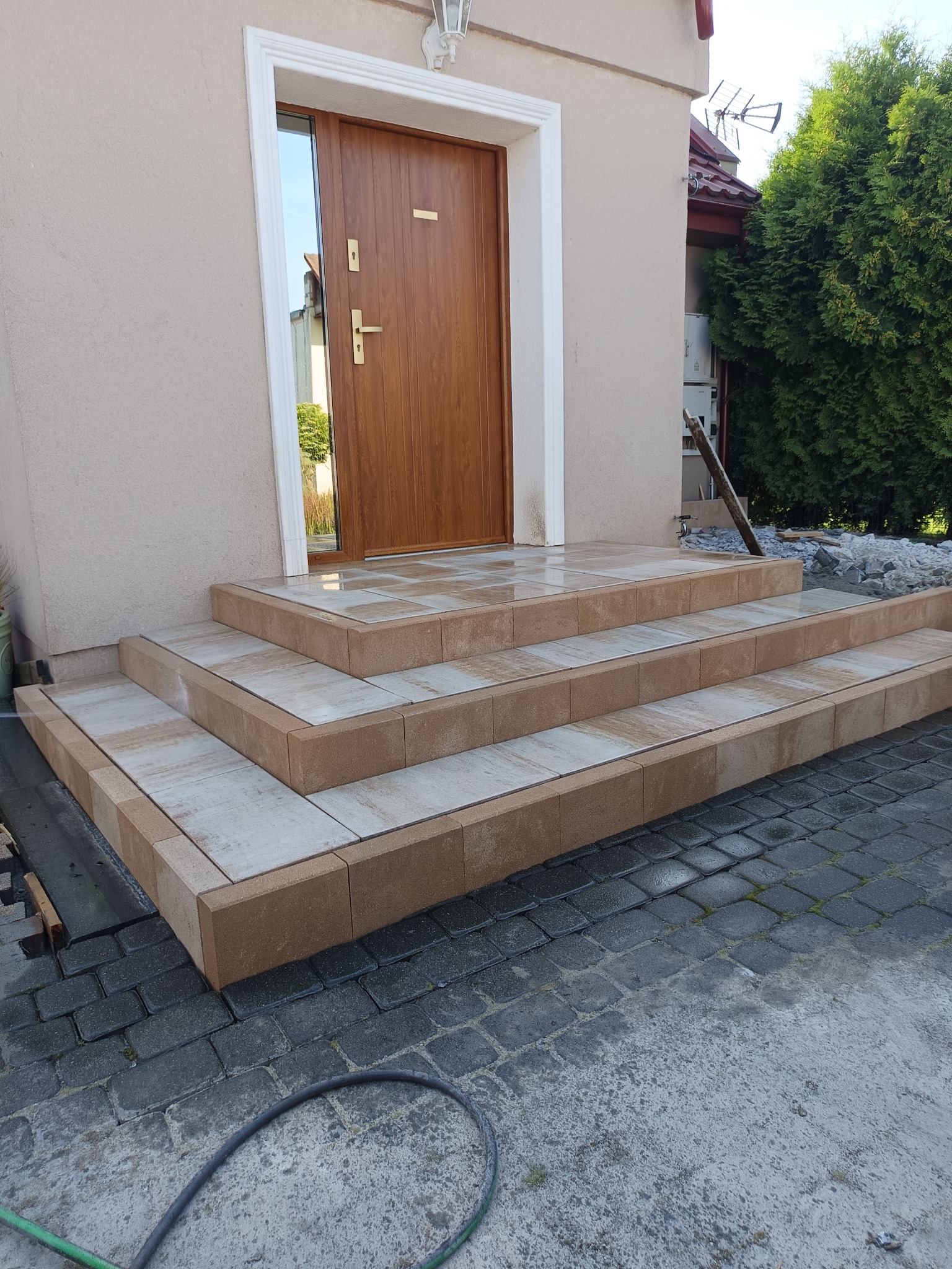 Nowe, trzypoziomowe schody z kostki brukowej prowadzące do drzwi wejściowych. Jasne płyty na stopniach, brązowe obrzeża. Częściowo ułożona kostka na podjeździe.