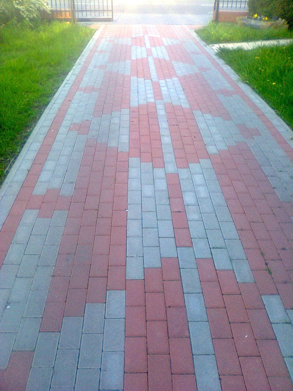 Ścieżka z kostki brukowej w odcieniach czerwieni i szarości, prowadząca do furtki. Wzór ułożenia kostki przypomina zygzak, po bokach trawnik. Ujęcie z dołu.