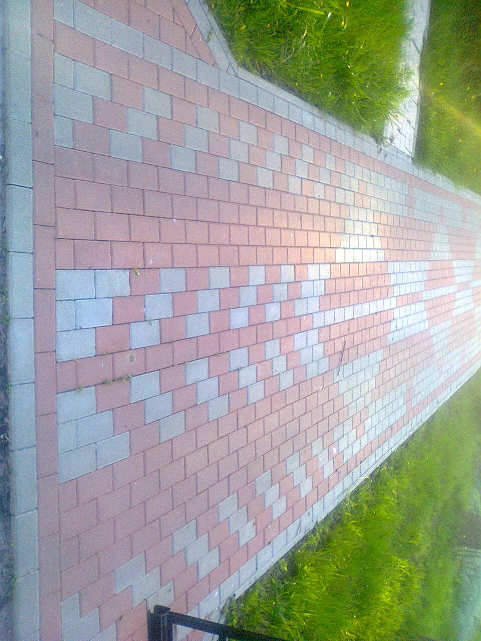 Ułożona kostka brukowa w geometryczny wzór, z widoczną zielenią trawnika po bokach. Czerwono-szara kolorystyka kostki. Widok z góry, fragment ogrodzenia w rogu.