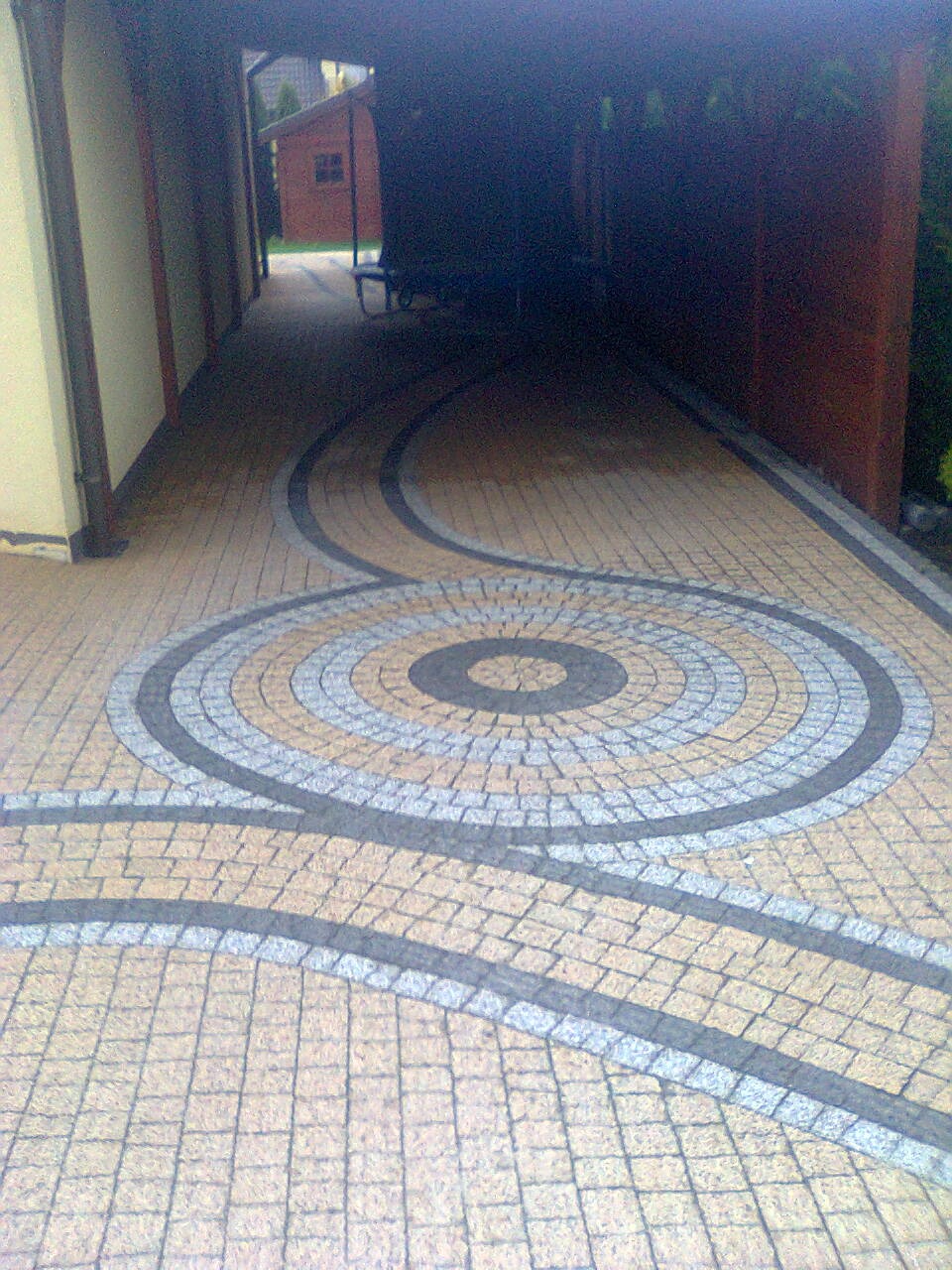 Ułożona kostka brukowa z wzorem okręgu i falistych linii w odcieniach beżu i szarości, prowadząca do zacienionego wjazdu.
