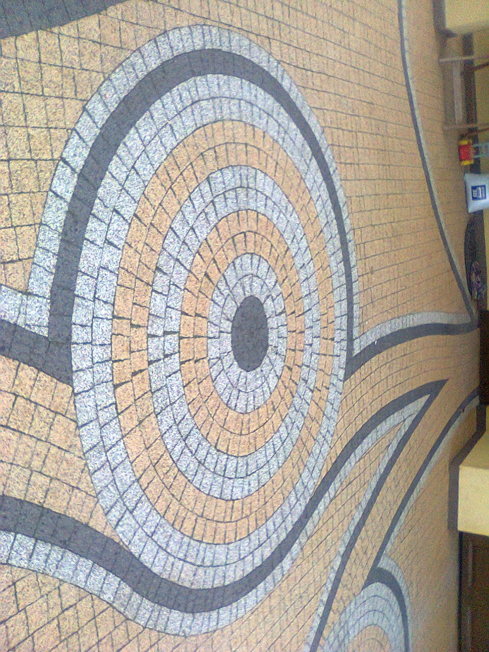 Mozaika z kostki granitowej w odcieniach beżu, szarości i czerni, tworząca wzór koncentrycznych okręgów i łuków. Nietypowy układ kostki, zbliżenie na detal.