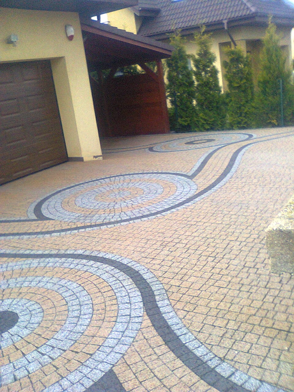 Efektowne ułożenie kostki brukowej w geometryczne wzory na podjeździe, z garażem i zielenią w tle. Widoczna precyzja wykonania i dbałość o detale.