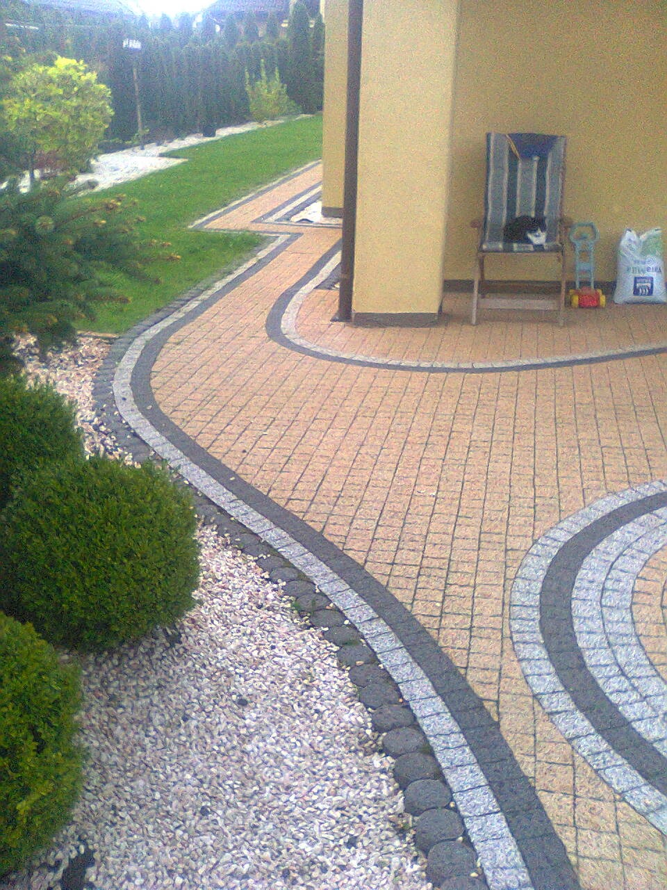 Kaskadowa ścieżka z kostki brukowej w odcieniach beżu i szarości, obrzeża z ciemnej kostki. W tle trawnik, krzewy i dom. Na krześle siedzi kot.