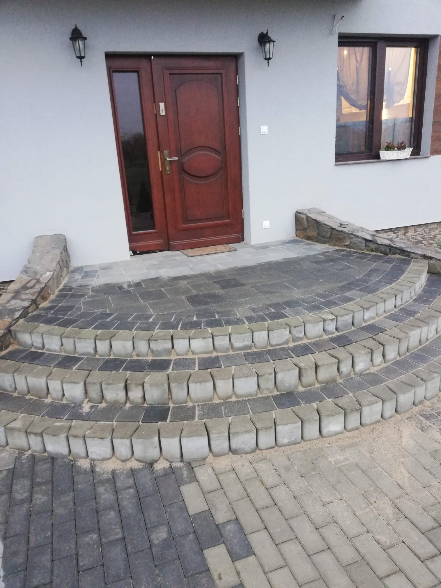 Schody wejściowe z kostki brukowej w odcieniach szarości, prowadzące do brązowych drzwi wejściowych w jasnym budynku. Widoczne lampy zewnętrzne i okno.
