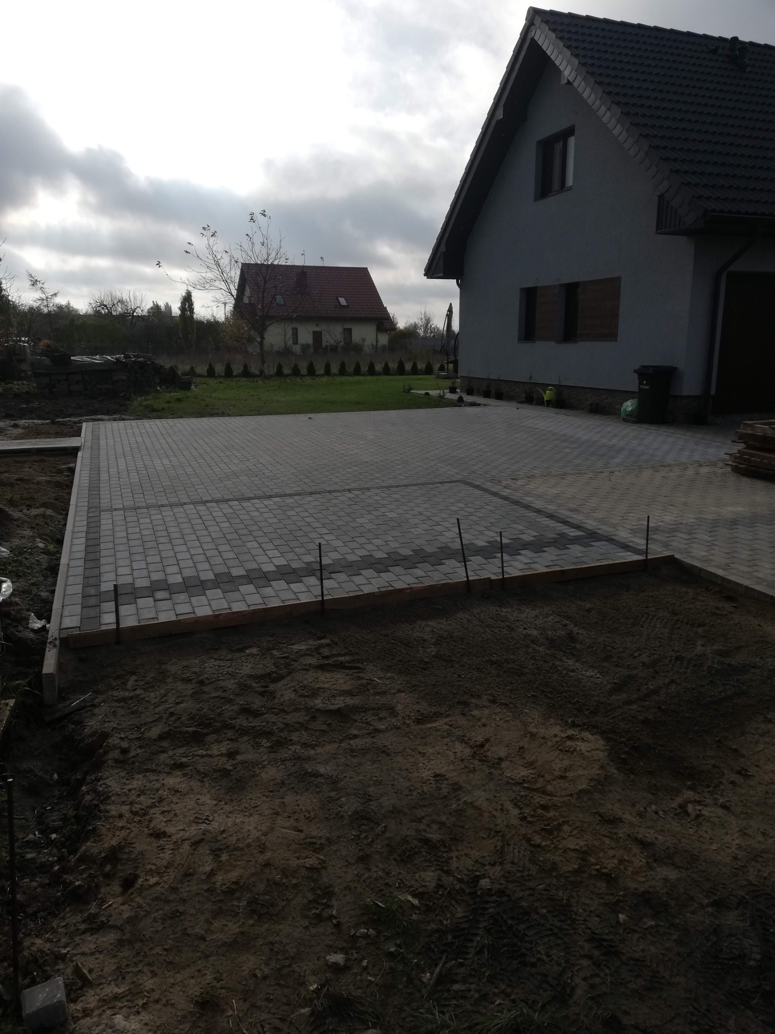 Częściowo ułożona, szara kostka brukowa na podjeździe przed domem w pochmurny dzień. Widoczne drewniane obramowanie i przygotowany teren pod dalsze prace.