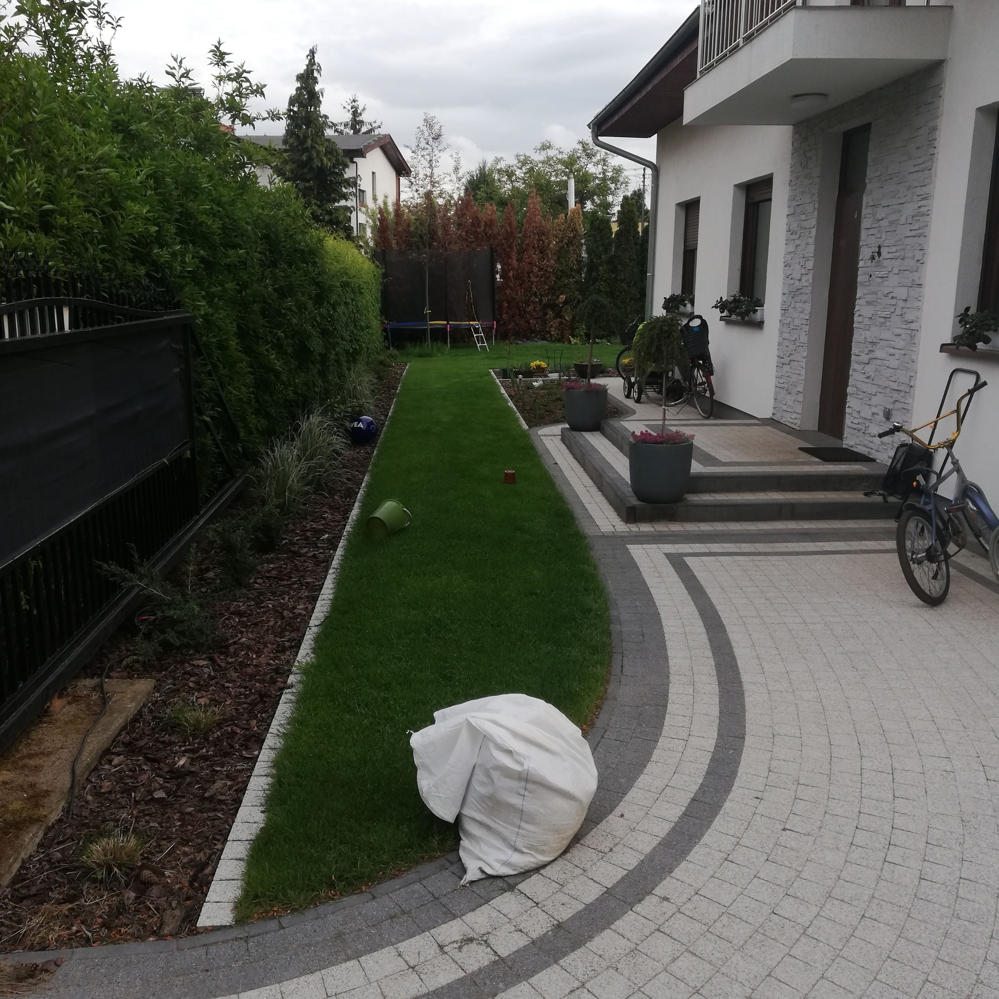 Ukończony chodnik z kostki brukowej w łuki, trawnik z rolki, rabata z korą i ogrodzenie panelowe przy domu z kamienną elewacją. Widok na podwórko.