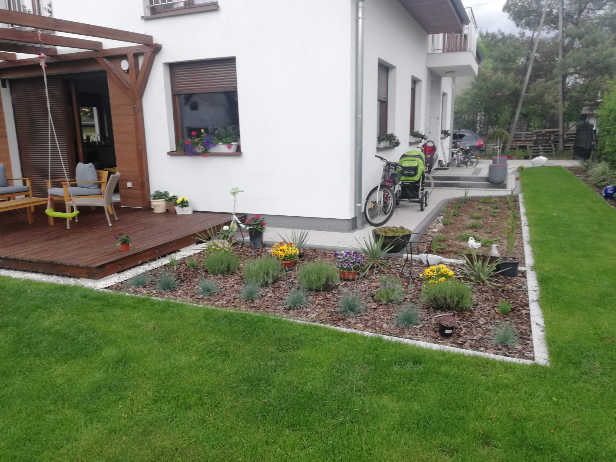 Estetyczny ogród przydomowy z drewnianym tarasem, rabatami kwiatowymi wykończonymi obrzeżem i równo przystrzyżonym trawnikiem.