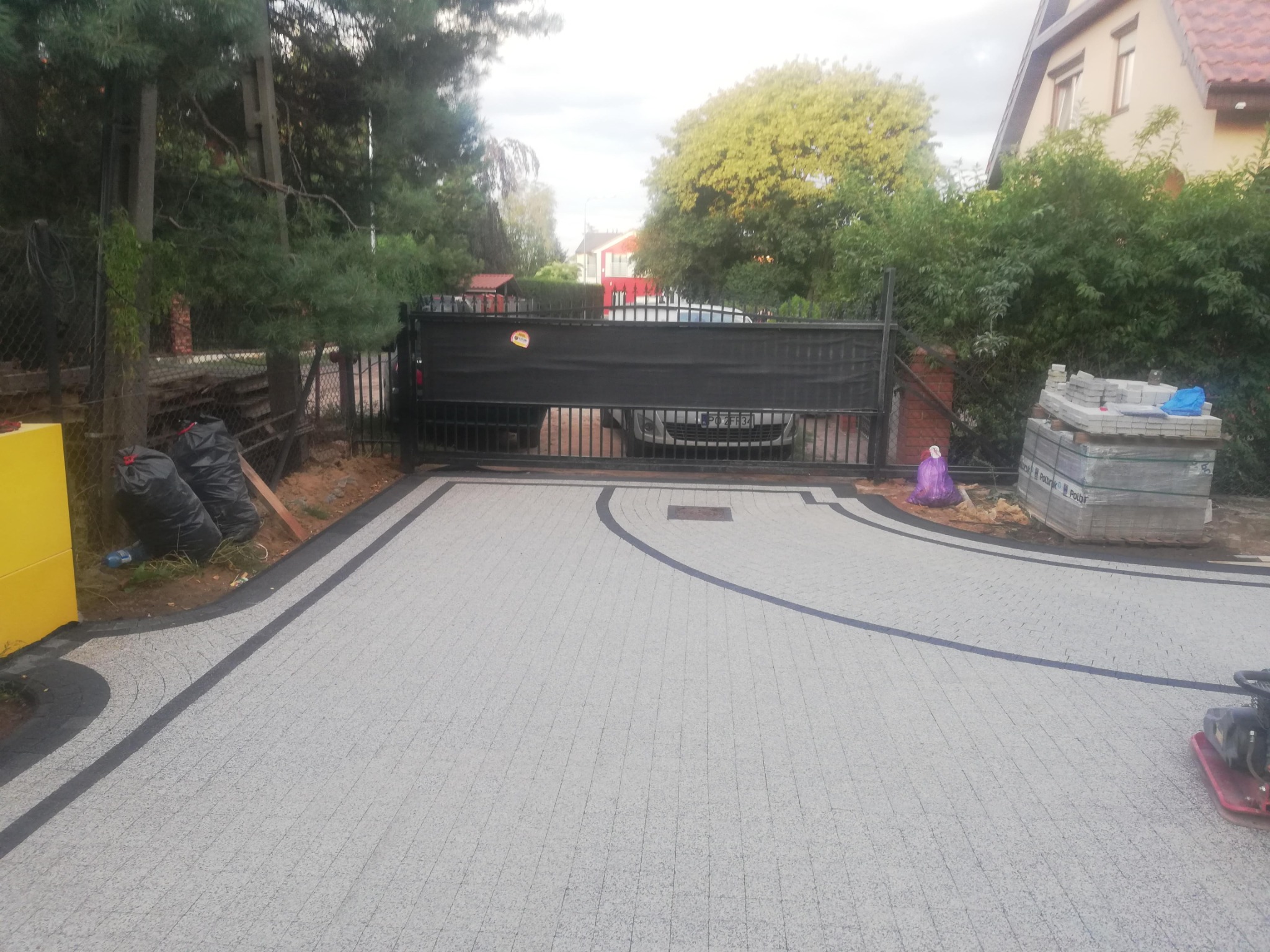 Ułożona szara kostka brukowa z ciemnymi obrzeżami na podjeździe, brama w tle i stos kostki obok. Widoczny zagęszczarka wibracyjna.