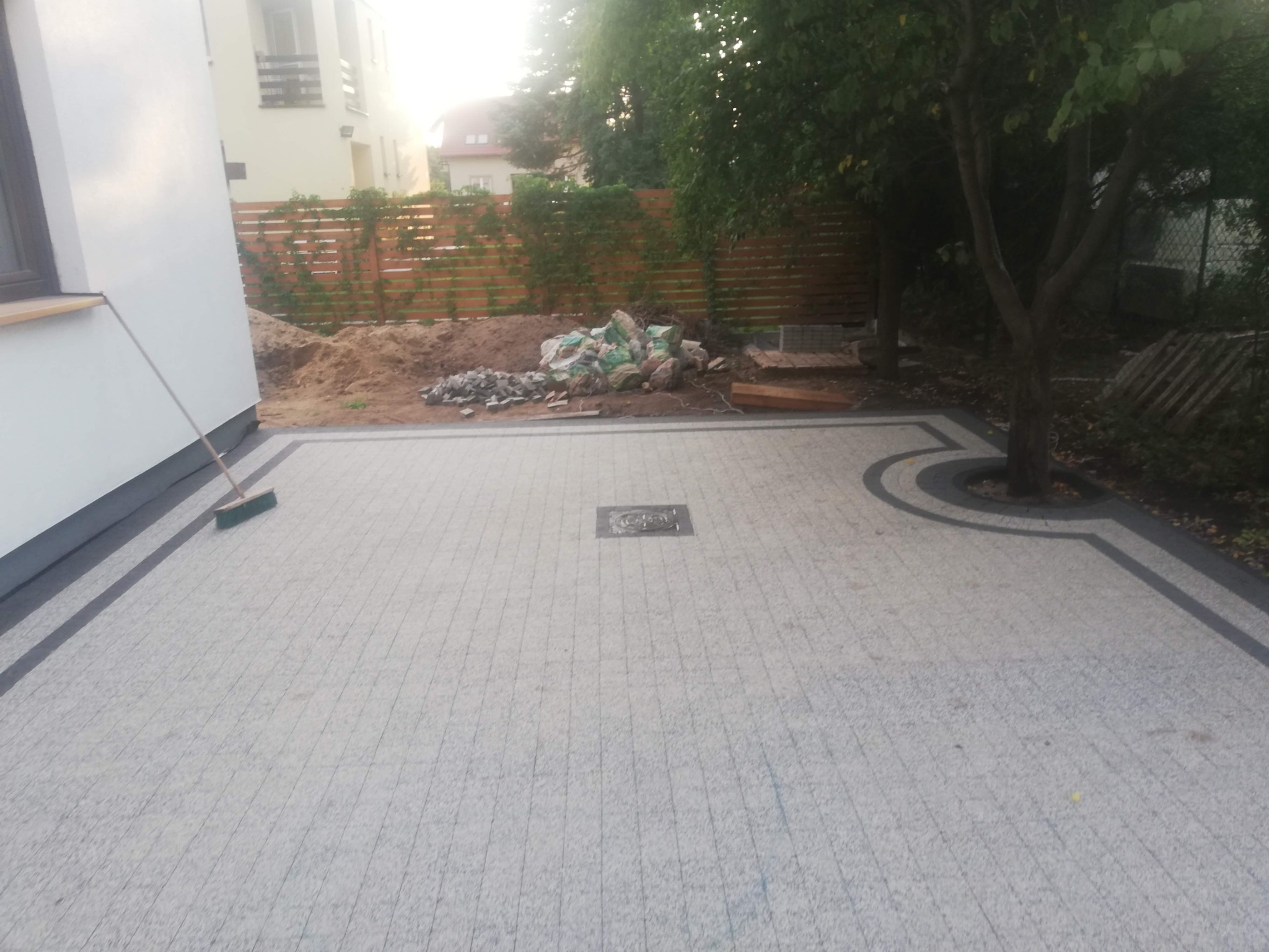 Wykończona nawierzchnia z jasnej kostki brukowej z ciemną obwódką, studzienka kanalizacyjna na środku. W tle widoczny teren przygotowany pod dalsze prace.