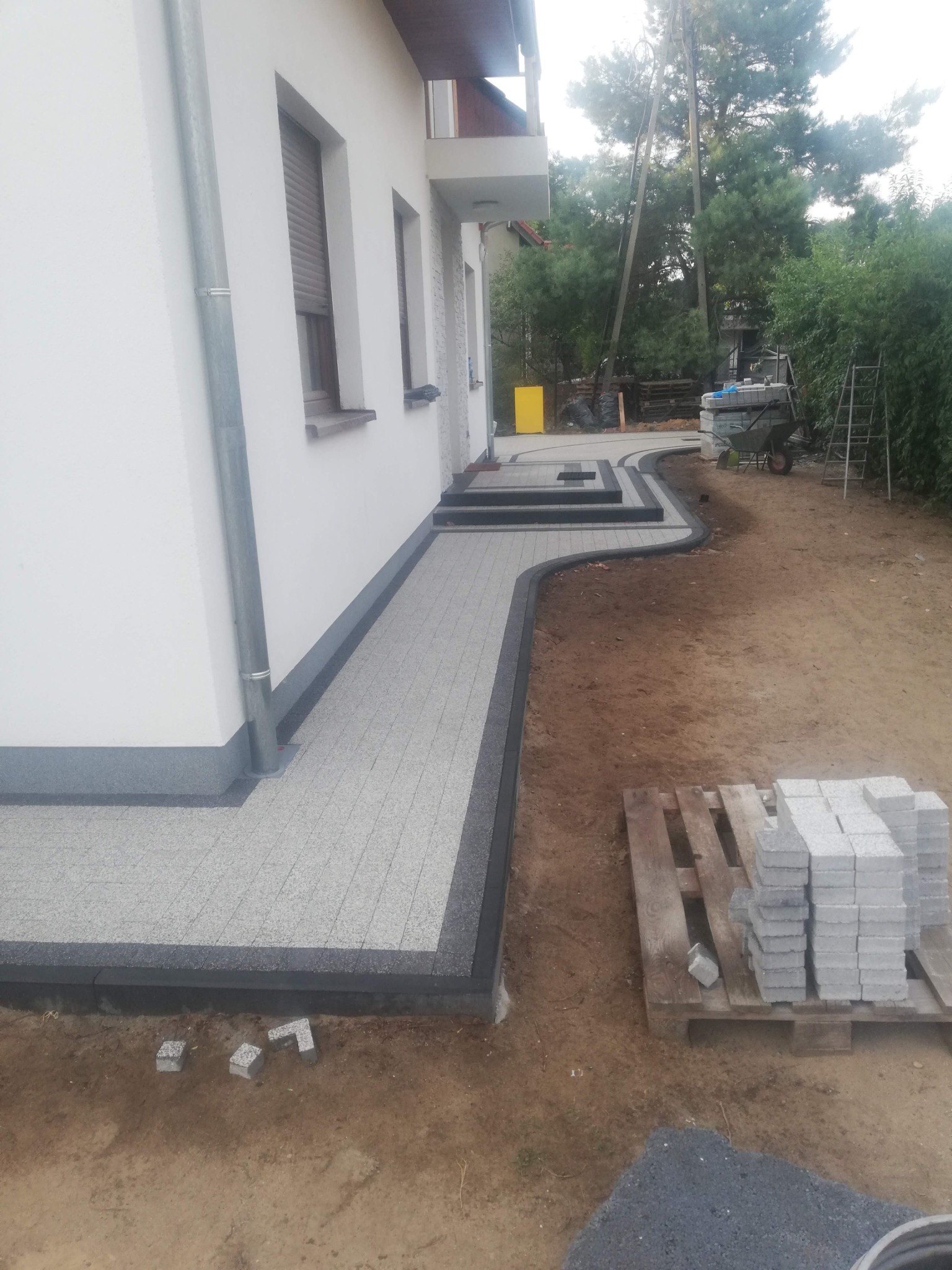 Ścieżka z kostki brukowej przy białym budynku, z eleganckim czarnym obramowaniem. Na palecie leżą kostki, a w tle widoczna taczka i drabina.