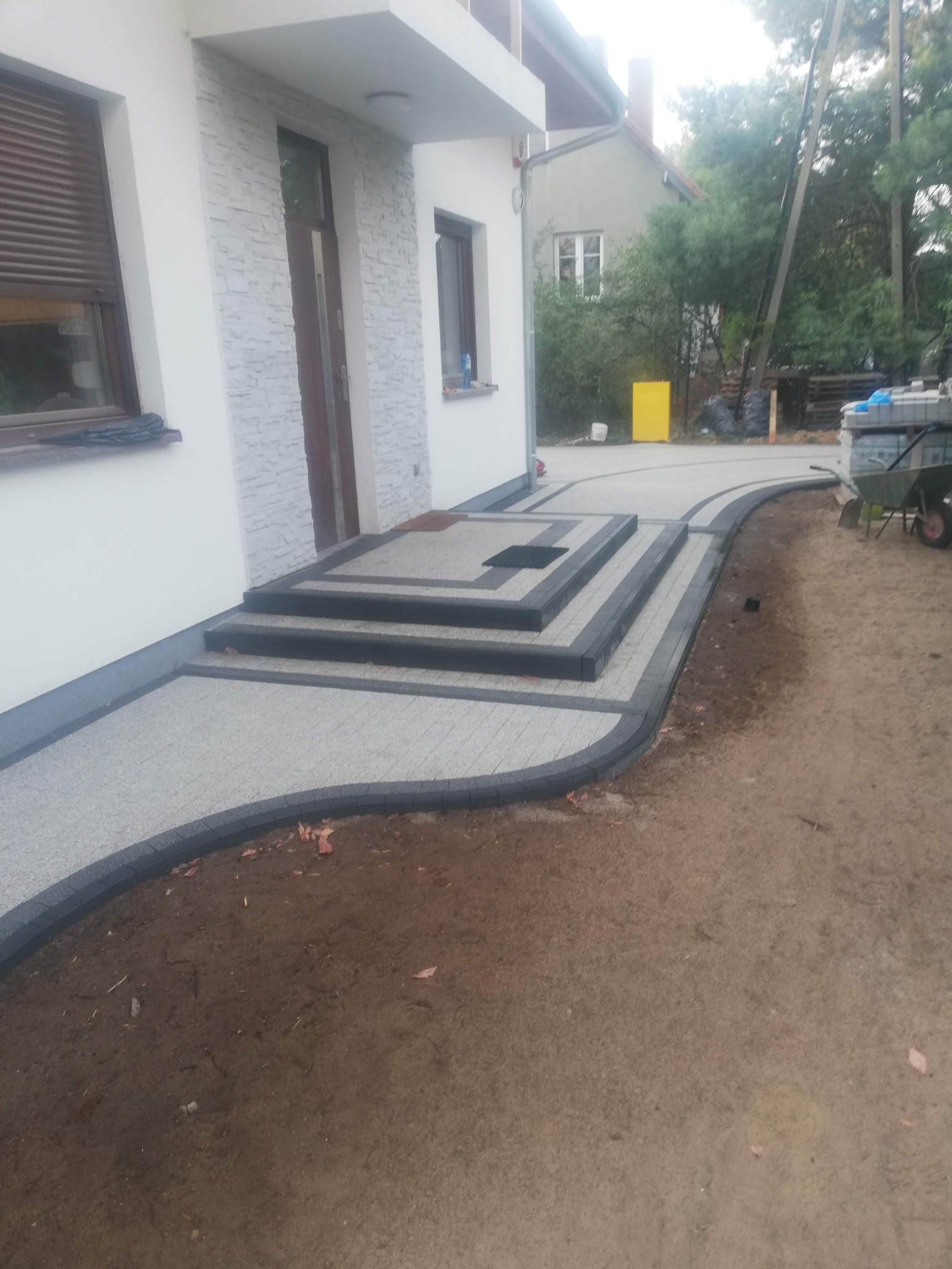 Ułożona kostka brukowa przed domem z trzema schodami, obrzeża w kolorze grafitowym. Widoczne elementy domu i fragment terenu.