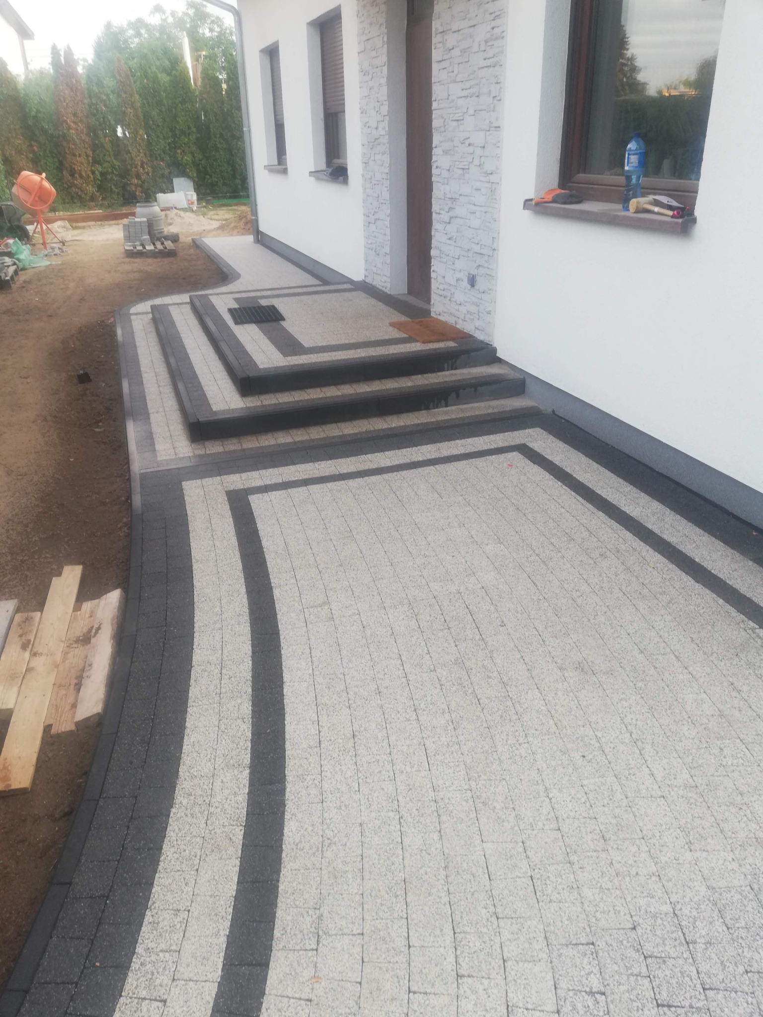 Ułożona kostka brukowa w dwóch kolorach, tworząca wzór na podjeździe i schodach wejściowych do domu. Widoczny mikser budowlany w tle, sugerujący prace wykończeniowe.