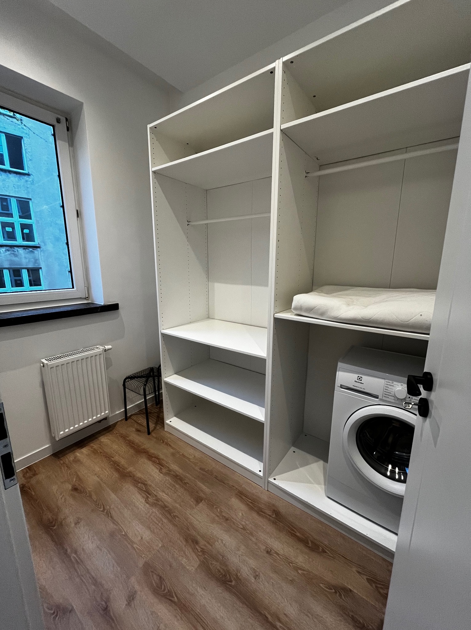 Biała zabudowa meblowa z pralką wnękową i półkami, widok z perspektywy wejścia, drewniana podłoga, okno z widokiem na budynek w tle.