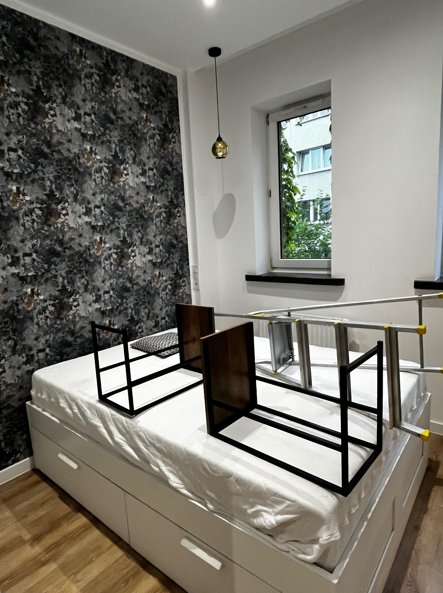 Pokój w trakcie przygotowań do sprzątania: rozłożona drabina, odstawione krzesła na łóżku z szufladami, wzorzysta tapeta na ścianie i okno z widokiem na zieleń.