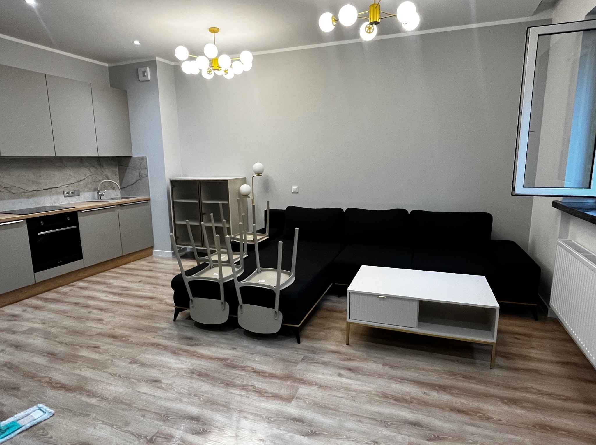 Wnętrze mieszkania po sprzątaniu: parkiet, narożnik, odwrócone krzesła, szafki kuchenne i mop na podłodze. Widok na wysprzątane mieszkanie z perspektywy podłogi.