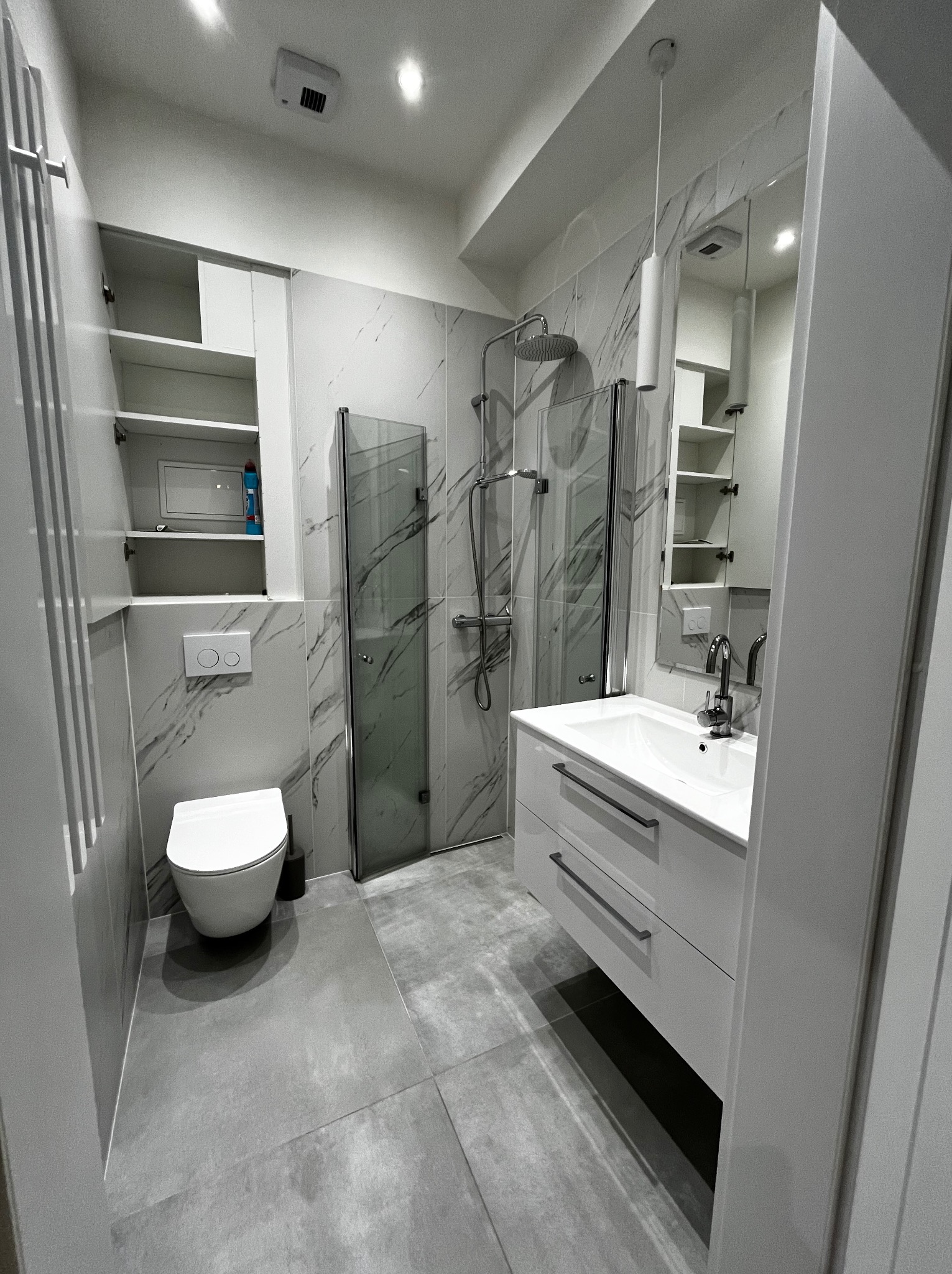 Wnętrze czystej, szaro-białej łazienki po sprzątaniu. Widoczna kabina prysznicowa, umywalka z szafką, toaleta i otwarte szafki z półkami. Marmurowe płytki na ścianach.