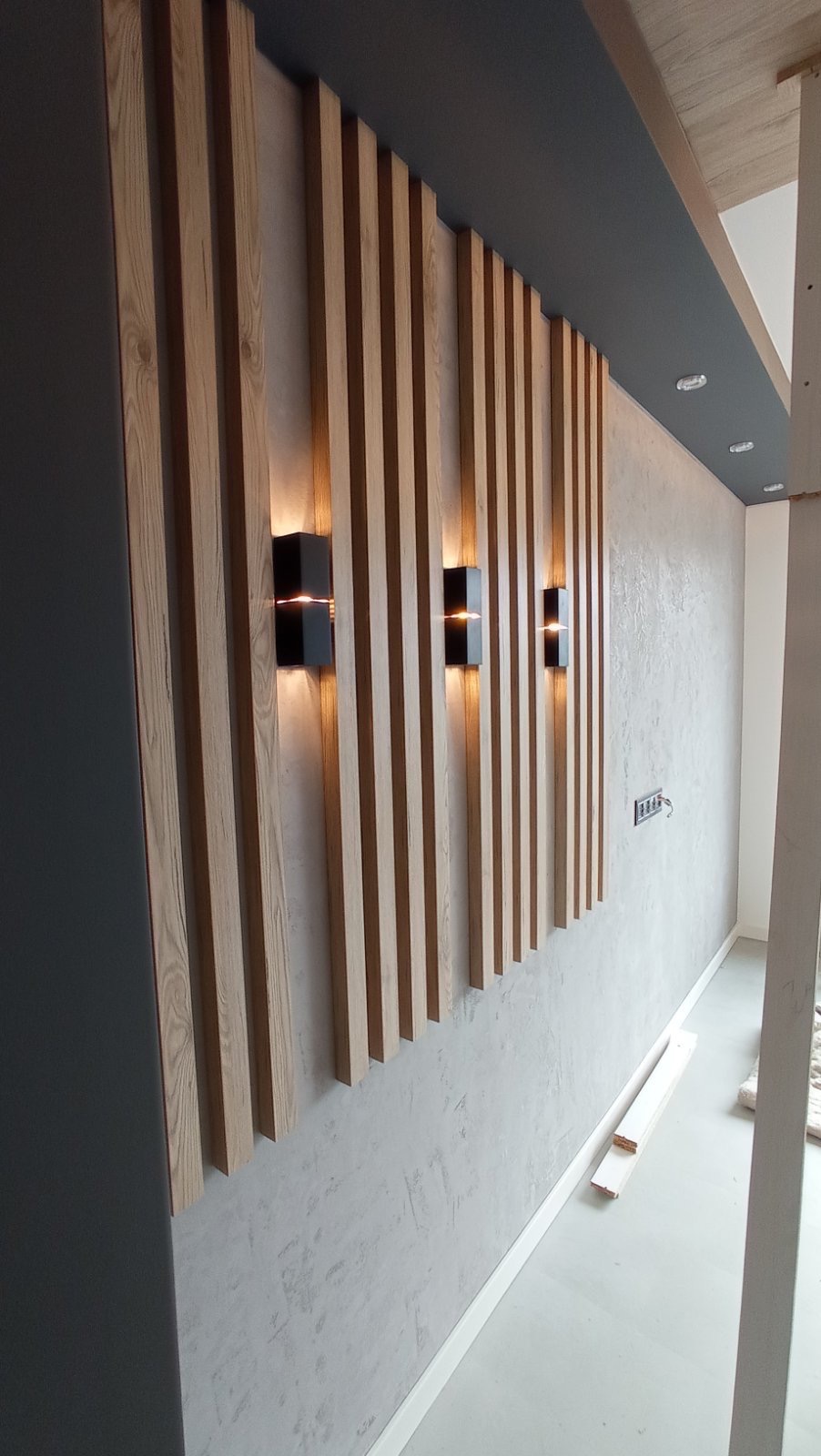Nowoczesna ściana z drewnianych paneli i oświetleniem punktowym. Szara ściana wykończona dekoracyjnym tynkiem. Widoczne listwy przypodłogowe i fragment sufitu z oświetleniem.
