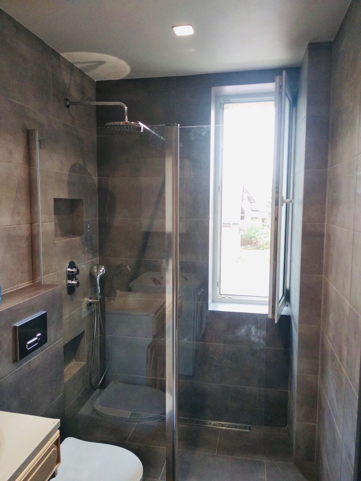 Nowoczesna kabina prysznicowa ze szklanymi drzwiami i deszczownicą w łazience wykończonej płytkami w odcieniach szarości. Widoczne okno i elementy armatury.