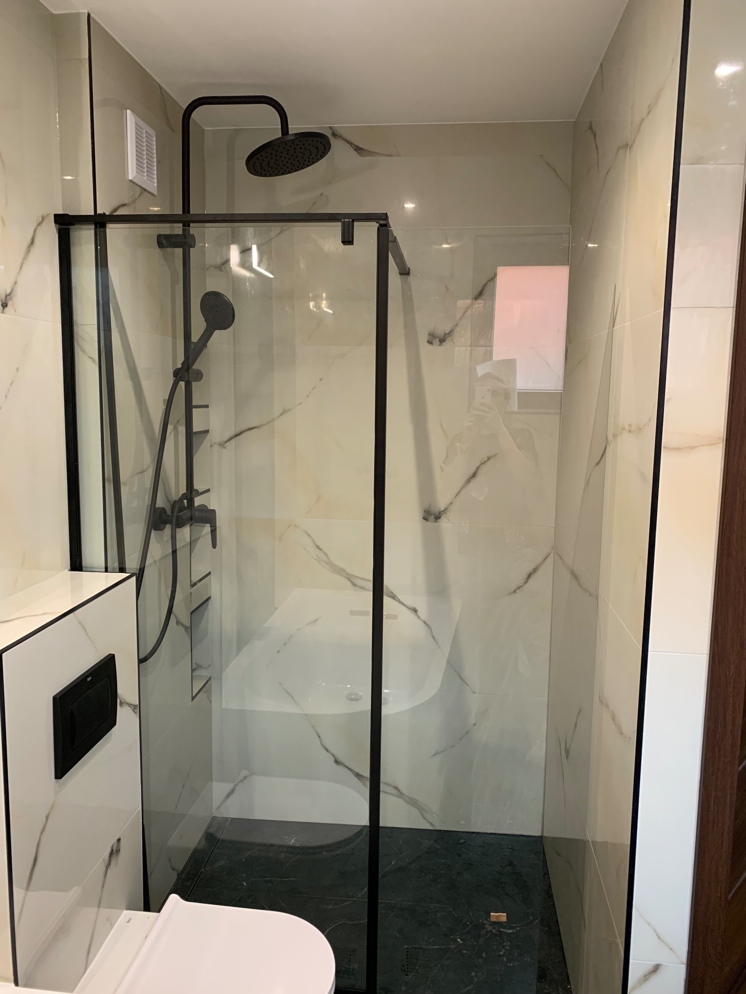 Nowoczesna kabina prysznicowa z czarnymi profilami i deszczownicą w wykończonej łazience z marmurowymi płytkami, widoczna toaleta i fragment drewnianego wykończenia.