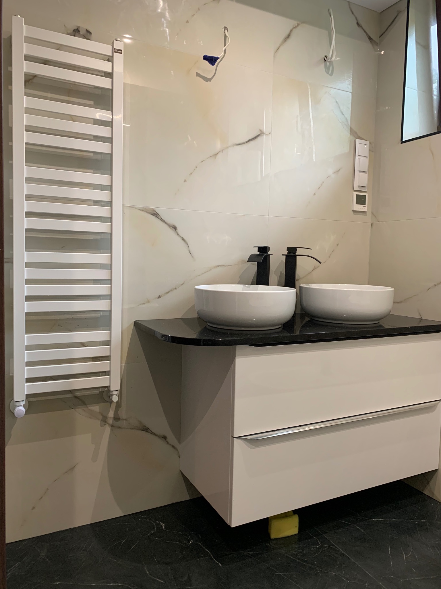 Nowoczesna łazienka z dwoma umywalkami na czarnym blacie, białą szafką i grzejnikiem drabinkowym na marmurowej ścianie. Widoczne niedokończone instalacje.