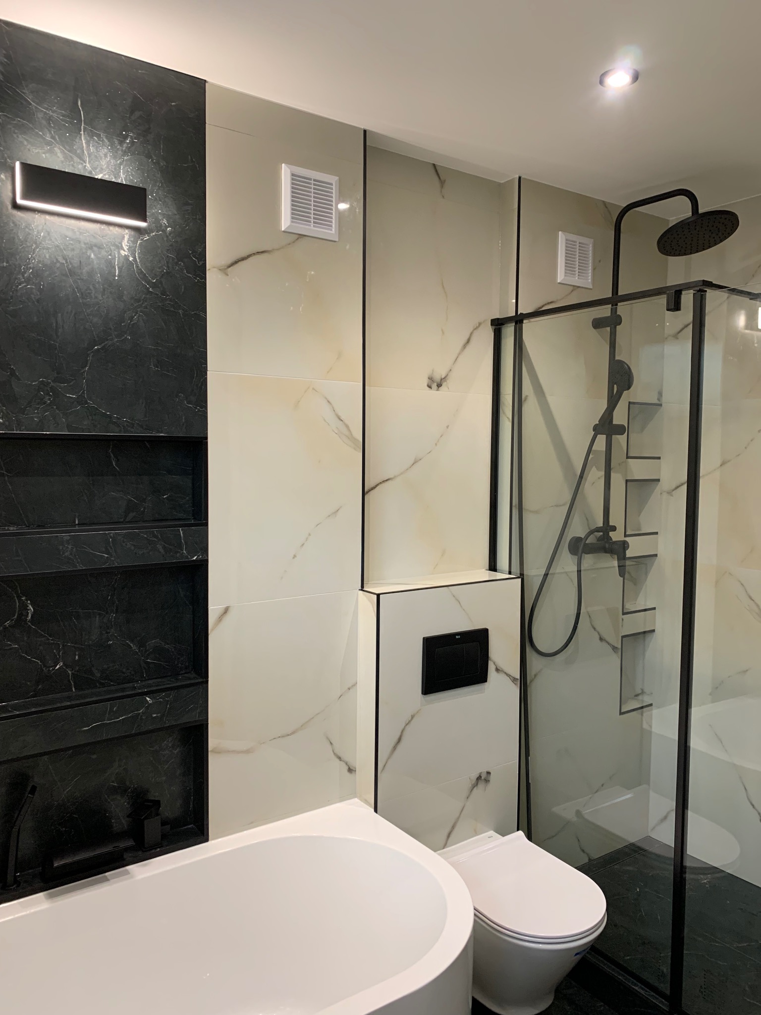 Elegancka łazienka w czerni i bieli z wanną, toaletą i kabiną prysznicową. Marmurowe płytki, czarne akcenty i oświetlenie tworzą nowoczesny wystrój.