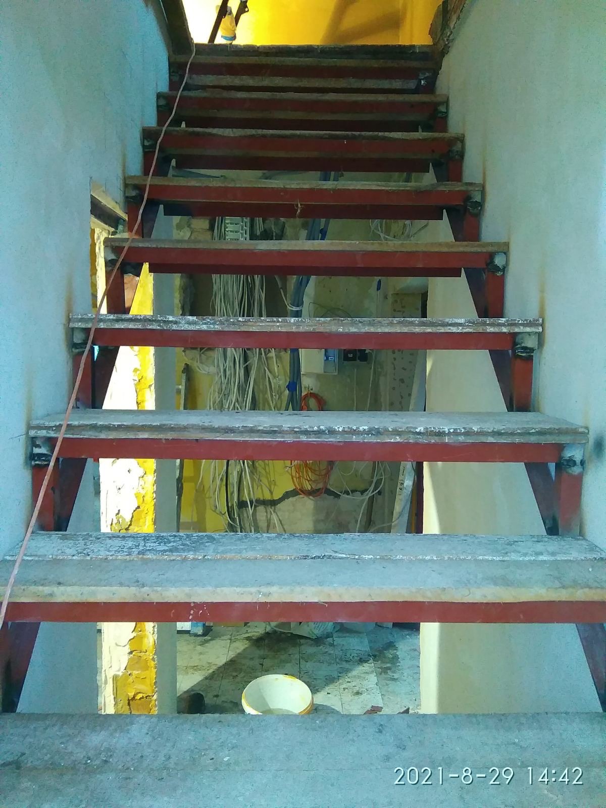 Metalowe schody w trakcie remontu, widoczne przewody elektryczne i fragmenty starej farby na ścianach. Ujęcie z dołu, podkreślające surowość wnętrza.