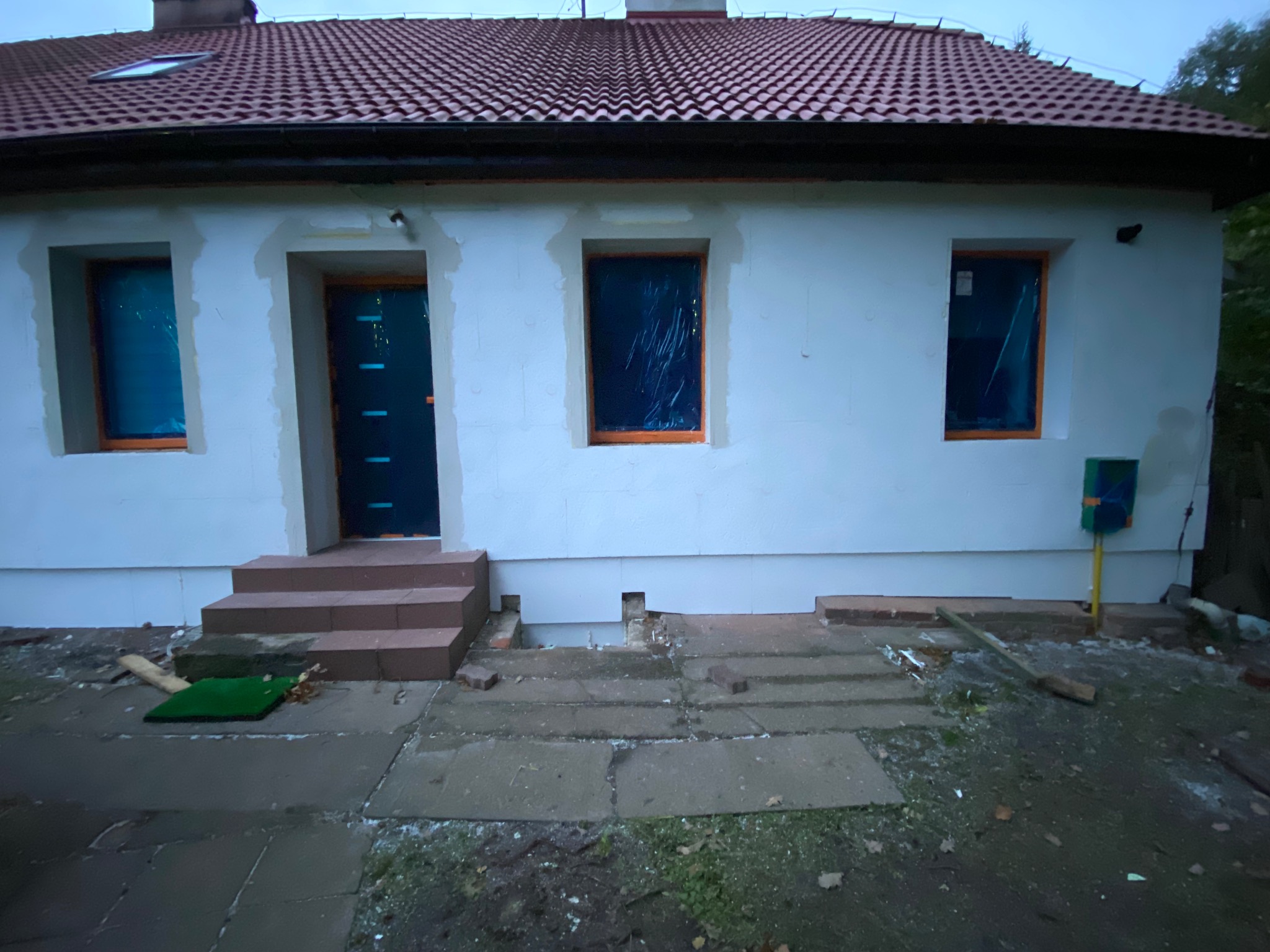 Dom jednorodzinny w trakcie prac remontowych: nowe okna zabezpieczone folią, otynkowane ściany, częściowo wykończone schody wejściowe. Widoczne ślady prac budowlanych.
