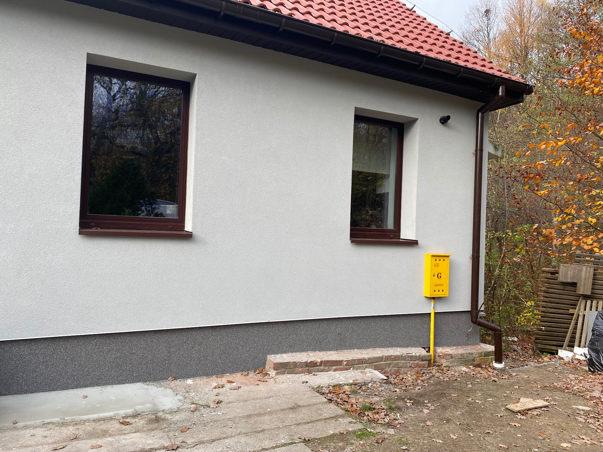 Elewacja domu z żółtą skrzynką gazową i brązowymi oknami. Widoczny fragment dachu z czerwoną dachówką i rynna. Jesienne liście na ziemi.