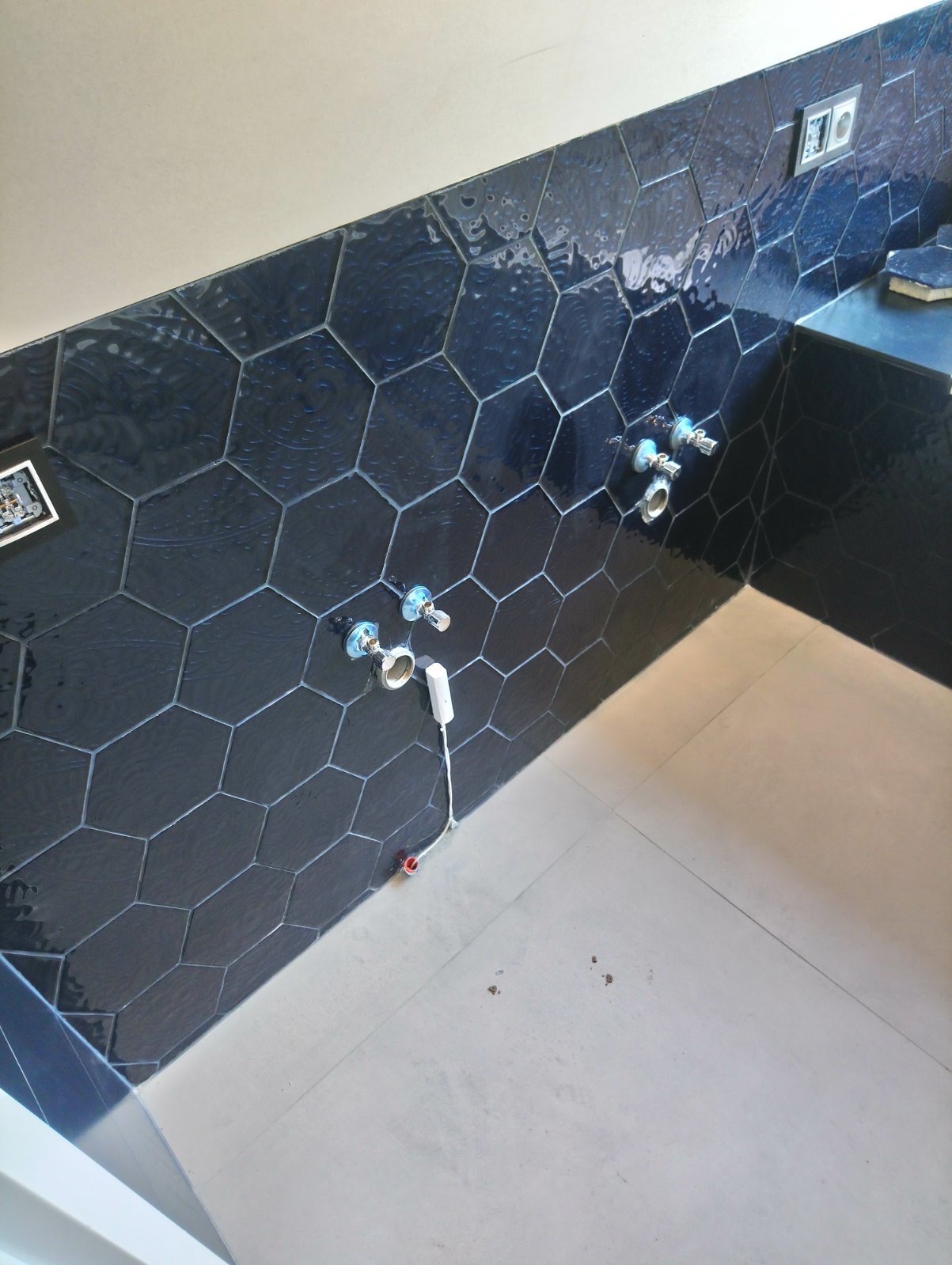 Łazienka w trakcie wykańczania: ciemne, heksagonalne płytki na ścianie, widoczne przyłącza wodne i fragment podłogi. Nieukończone wykończenie.