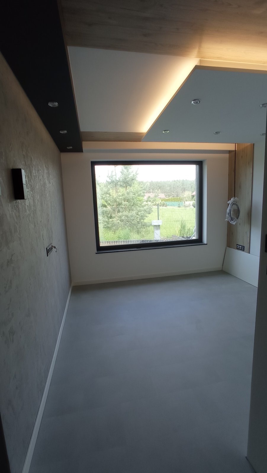 Nowoczesne wykończenie wnętrza: szara ściana z fakturą, gładka podłoga, okno z widokiem na zieleń, podwieszany sufit z oświetleniem LED. Minimalistyczny design.
