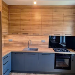 Nowoczesna kuchnia z drewnianymi górnymi szafkami i szarymi dolnymi, czarnymi uchwytami. Blat drewnopodobny, czarny panel nad kuchenką. Widoczny zlew i płyta gazowa.