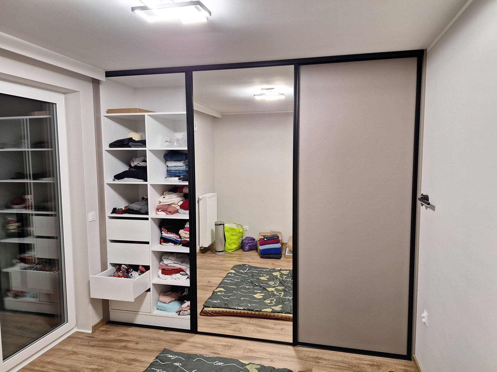 Szafa wnękowa z przesuwnymi drzwiami i lustrem, z otwartą częścią z półkami i szufladami, wypełniona ubraniami. Widok na wnętrze szafy odbity w lustrze.