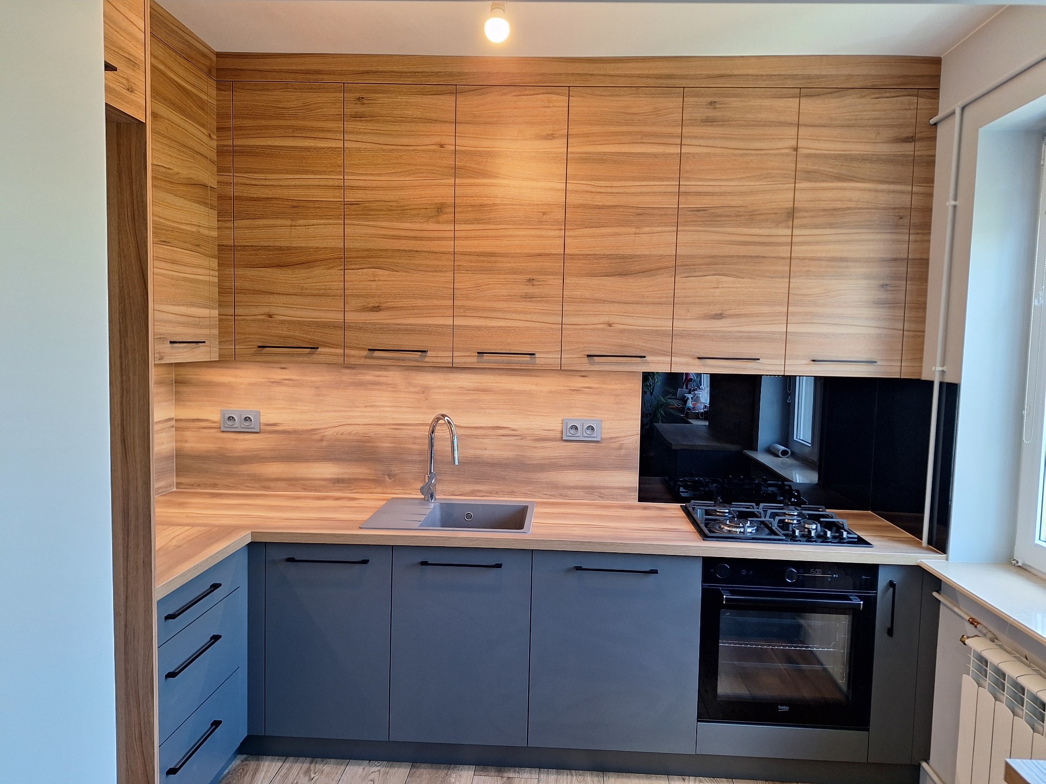 Nowoczesna kuchnia z drewnianymi górnymi szafkami i szarymi dolnymi, czarnymi uchwytami. Blat drewnopodobny, czarny panel nad kuchenką. Widoczny zlew i płyta gazowa.