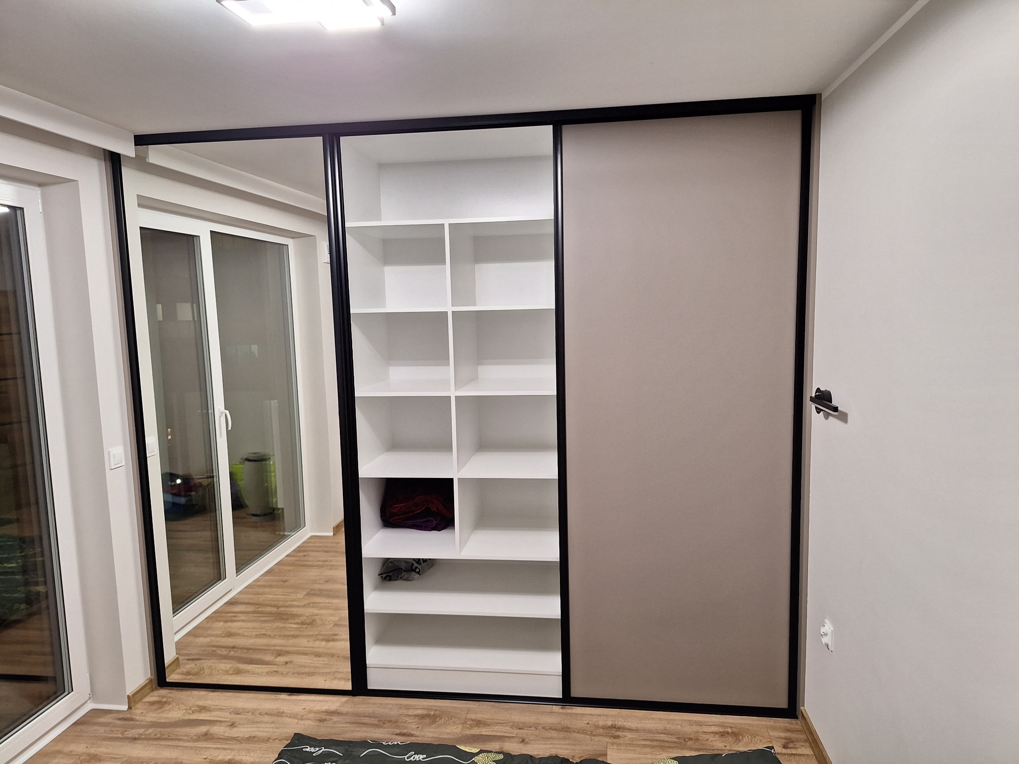 Szafa wnękowa z przesuwnymi drzwiami: lustro, beżowa płyta. Wnętrze z białymi półkami, widoczne ubrania. Czarna rama drzwi. Parkiet na podłodze.